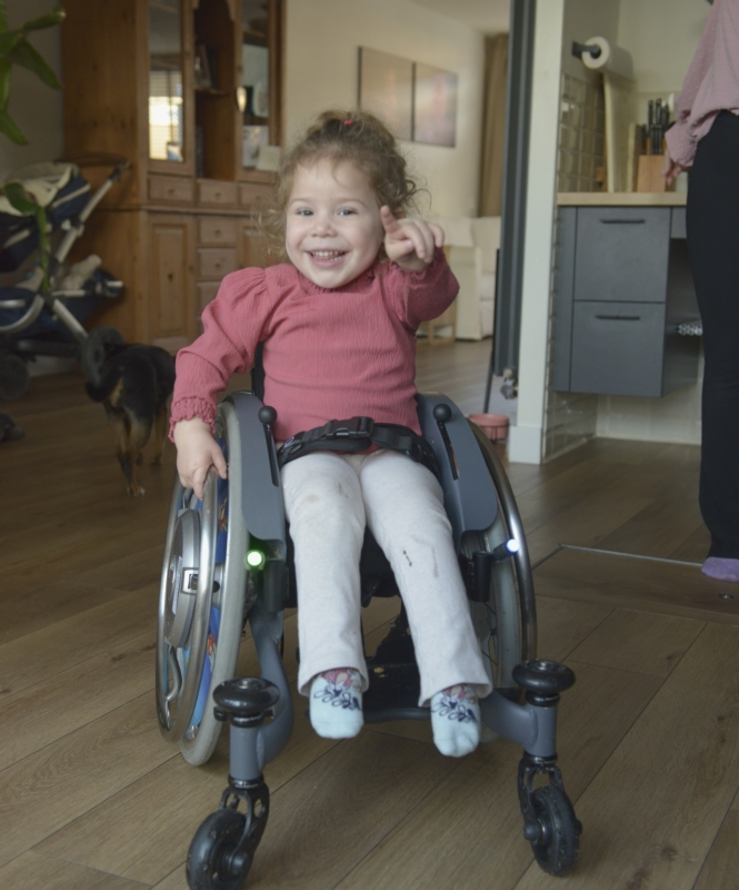 Kind Elena in rolstoel. Wijzend naar de camera.